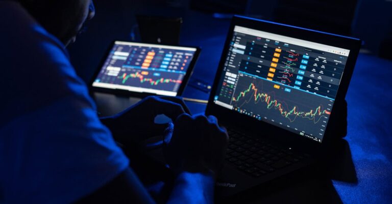 robo-advisers ai - Man analyzing financial charts and data on laptops in a dimly lit room, highlighting forex trading.