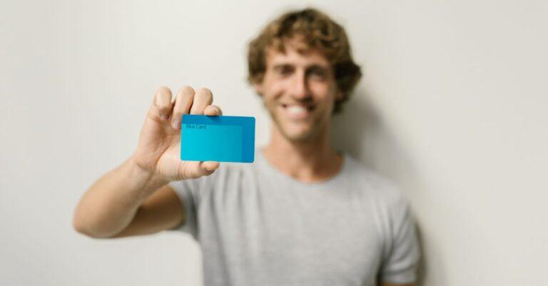 meta backs india - A smiling man in a gray shirt holds a blue card, offering a bright and friendly demeanor against a neutral