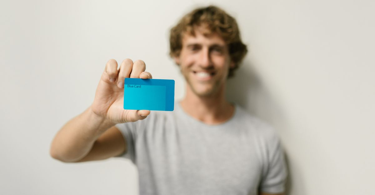 meta backs india - A smiling man in a gray shirt holds a blue card, offering a bright and friendly demeanor against a neutral