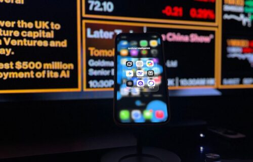 ai investment bust - Smartphone displaying AI apps in front of a financial data screen in London.
