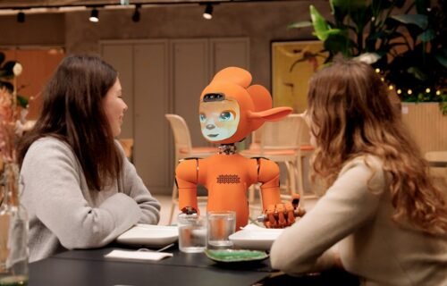 generative ai banking - Two women talk to an orange robot at a table.