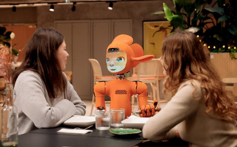 generative ai banking - Two women talk to an orange robot at a table.