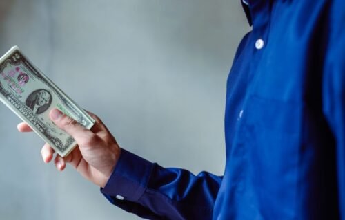 ai spending earnings - A person in a blue shirt holding a stack of US dollar bills, symbolizing wealth and finance.