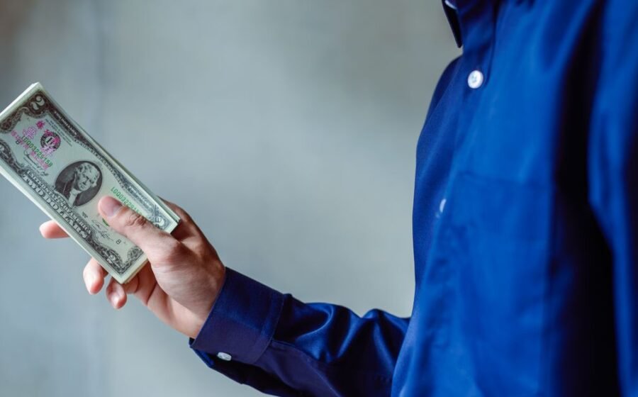 ai spending earnings - A person in a blue shirt holding a stack of US dollar bills, symbolizing wealth and finance.